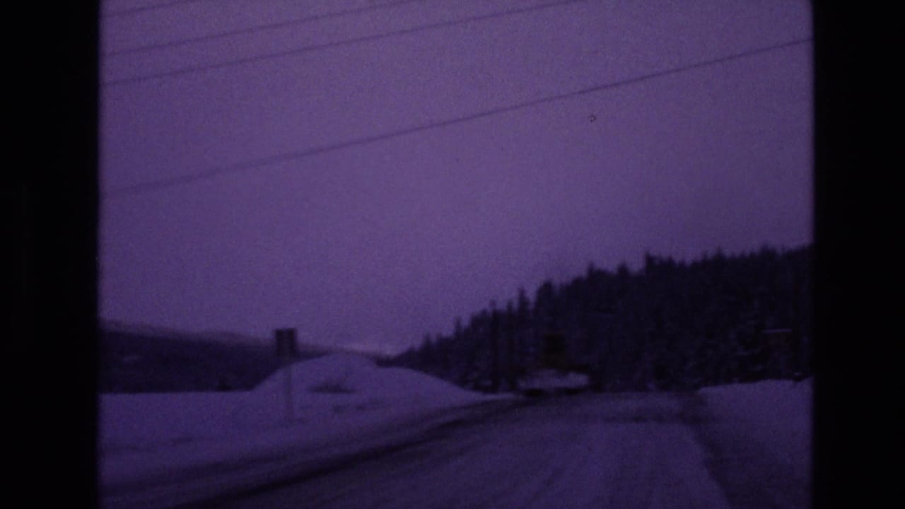 1970s Vail-area winter road view from a moving vehicle, with snowbanks, evergreen trees, utility lines, and a purple-toned dusk sky.