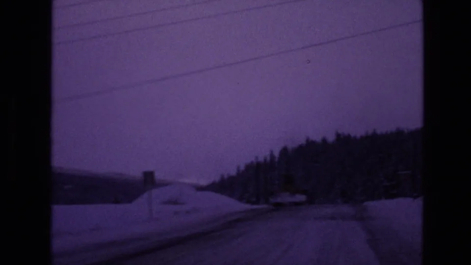 1970s Vail-area winter road view from a moving vehicle, with snowbanks, evergreen trees, utility lines, and a purple-toned dusk sky.