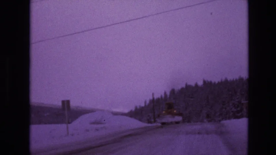 Moving 8mm view of a snowy Vail, Colorado roadside, showing a roadside sign, utility line, evergreen trees, and a distant winter hillside.