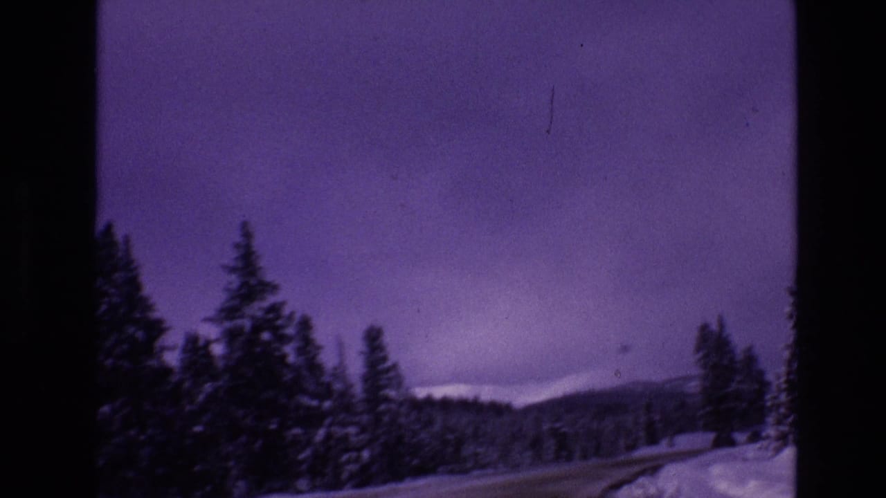 1974 8mm Vail, Colorado home-movie frame showing a snow-covered road curving through dark evergreen trees with low mountains beneath a purple winter sky.
