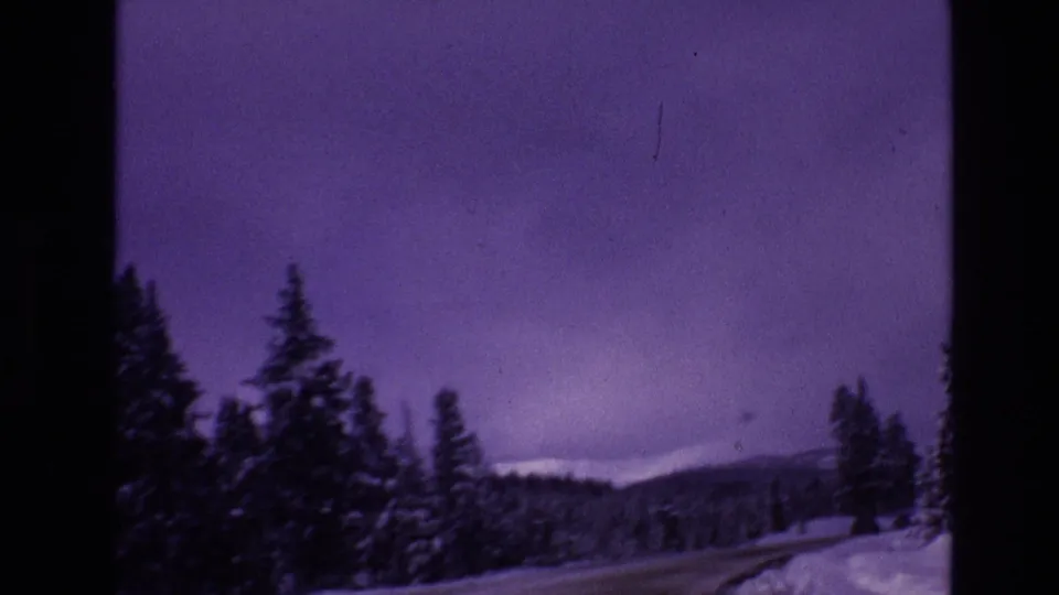 1974 8mm Vail, Colorado home-movie frame showing a snow-covered road curving through dark evergreen trees with low mountains beneath a purple winter sky.