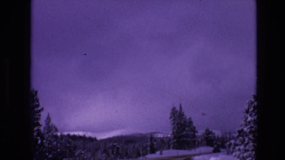1974 8mm Vail, Colorado home-movie frame with a snowy forest edge, low mountain ridge, and wide purple sky seen from a moving roadside viewpoint.