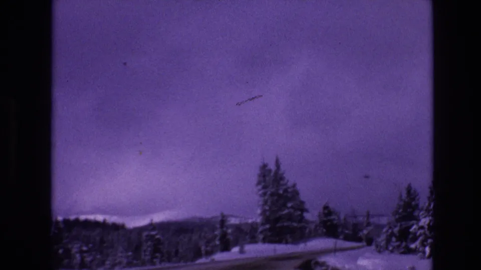 1974 8mm Vail, Colorado frame showing a snowy roadside curve, clustered evergreens, and distant winter hills under a muted purple dusk sky.