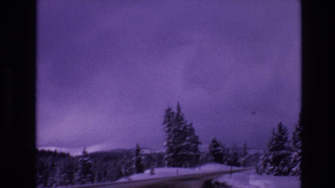 1974 Vail, Colorado 8mm frame showing a snow-covered road, roadside conifers, distant mountains, and an overcast purple-shifted sky.