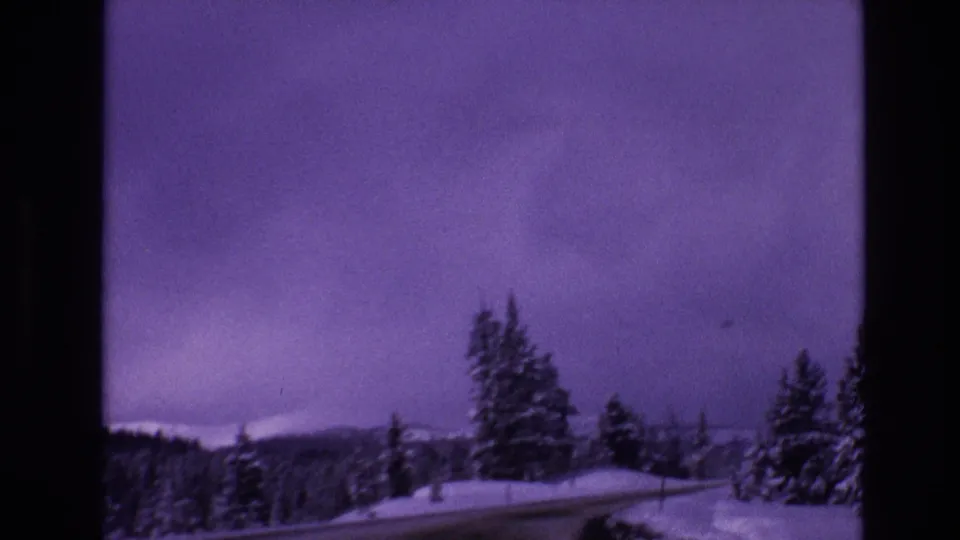 1974 Vail, Colorado 8mm frame showing a snow-covered road, roadside conifers, distant mountains, and an overcast purple-shifted sky.