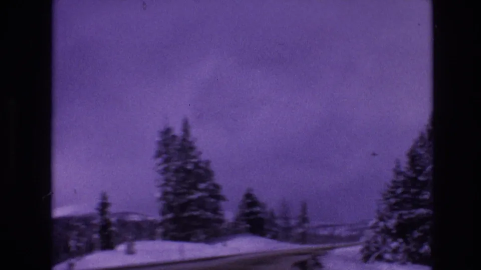 Soft 8mm frame from Vail, Colorado in 1974 showing snowbanks, conifers, a road edge, distant hills, and a magenta-purple winter sky.