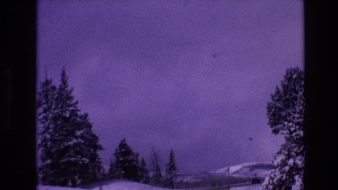 1974 Vail, Colorado 8mm frame showing a snow-covered roadside, dark evergreen trees, low fencing, and distant winter hills with a purple color cast.