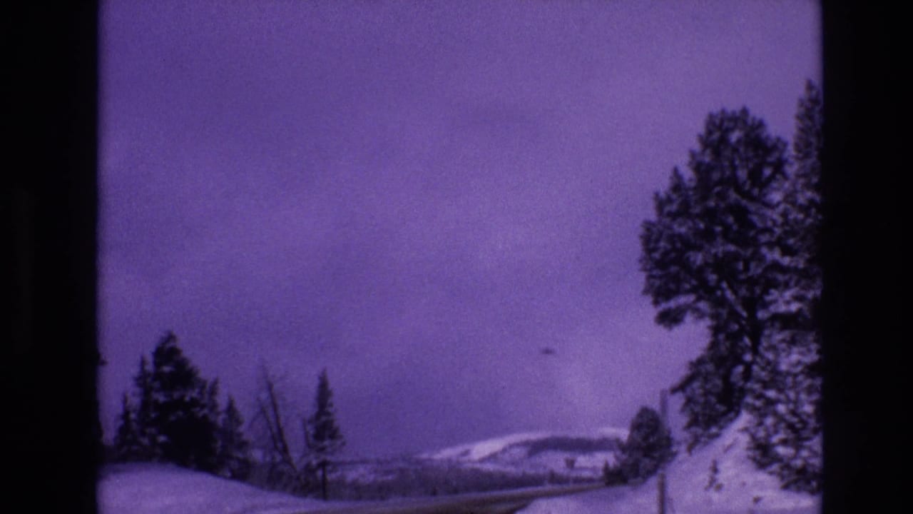 1974 Vail, Colorado home-movie frame looking past snowbanks and roadside trees toward pale hills, with dark frame borders and faded purple film color.