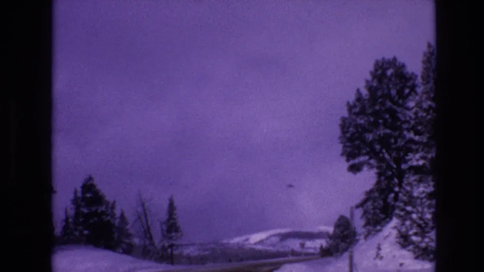 1974 Vail, Colorado home-movie frame looking past snowbanks and roadside trees toward pale hills, with dark frame borders and faded purple film color.