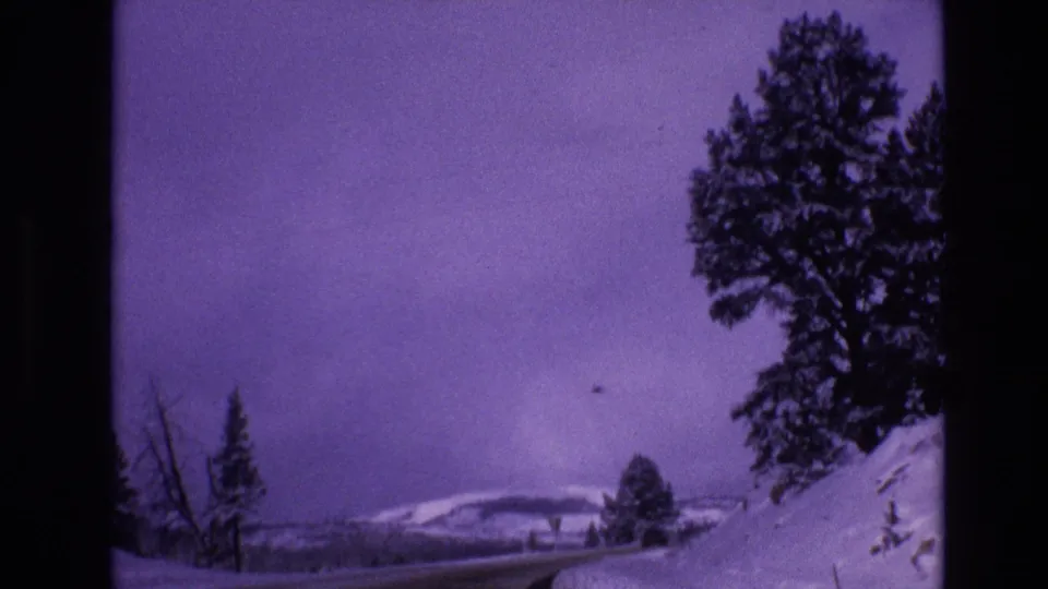 1974 Vail, Colorado 8mm frame with a tall evergreen rising from a snowbank, a faint road or fence line, and distant snowy hills under a faded purple cast.
