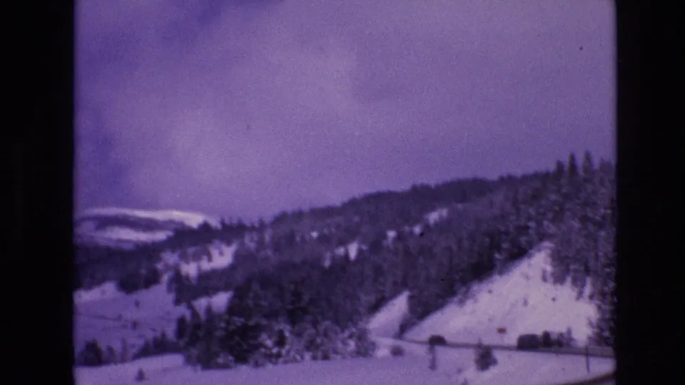 1974 Vail, Colorado home-movie frame showing a snowy roadside bend, forested hillside, low fence, and winter mountain terrain with soft 8mm focus.