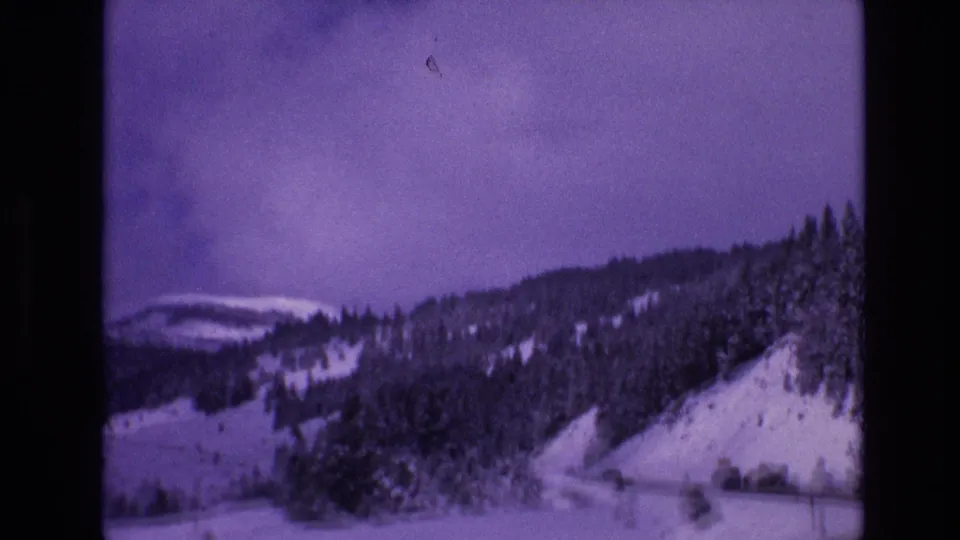 Vail, Colorado 1974 8mm frame showing a snowy evergreen-covered hillside with a road running along the mountain slope under an overcast sky.