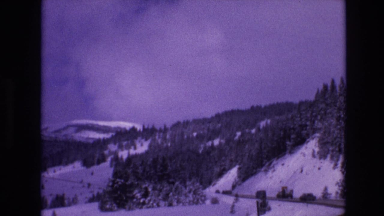 Snow-covered mountain road with cars along the hillside below evergreen trees.