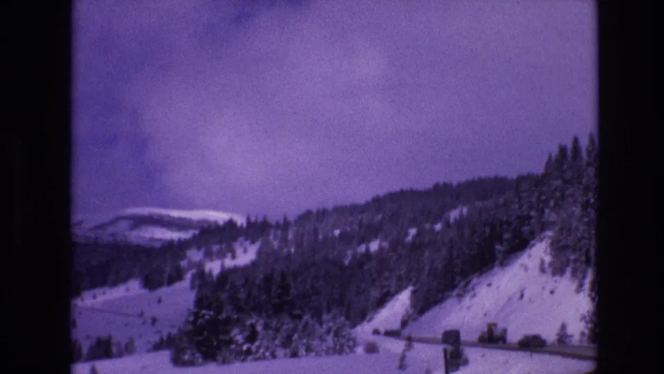 Vail, Colorado 1974 frame with small cars on a road cut into a snowy mountain slope below dense evergreen trees and distant ridgelines.