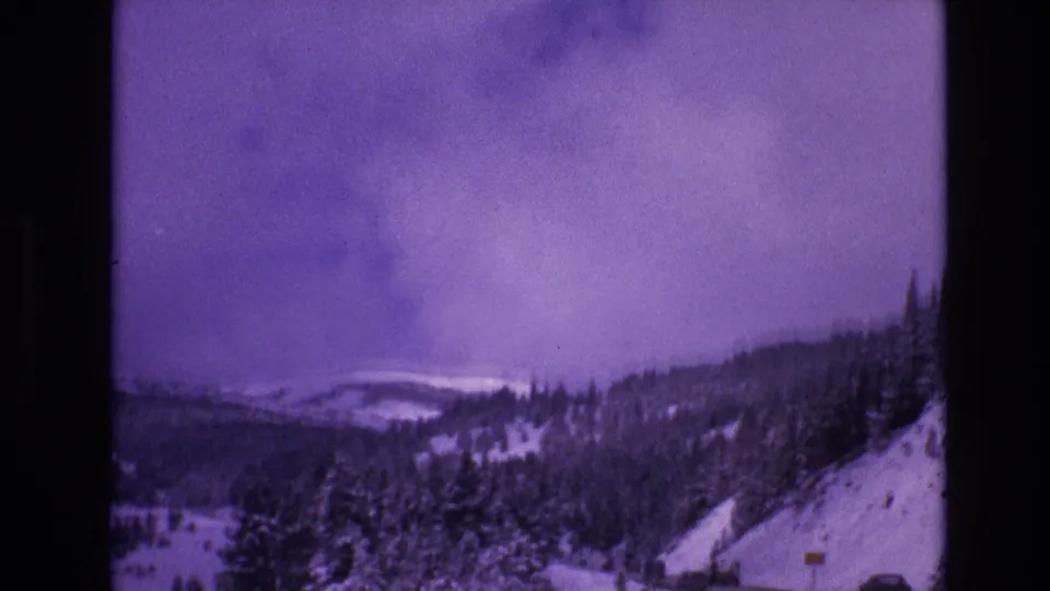 1974 8mm home-movie frame from Vail, Colorado showing snow-covered forest slopes and distant ridges beneath a heavy purple-tinged sky.