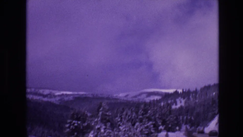 1974 8mm frame from Vail, Colorado looking across a snowy roadside valley toward forested hills and pale winter ridges under a clouded sky.