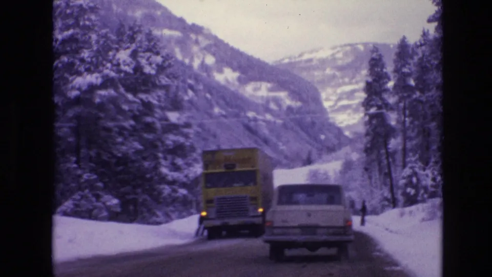 1974 Vail, Colorado road scene with a yellow semi-truck, a white station wagon, snow-laden trees, and mountains in the background.