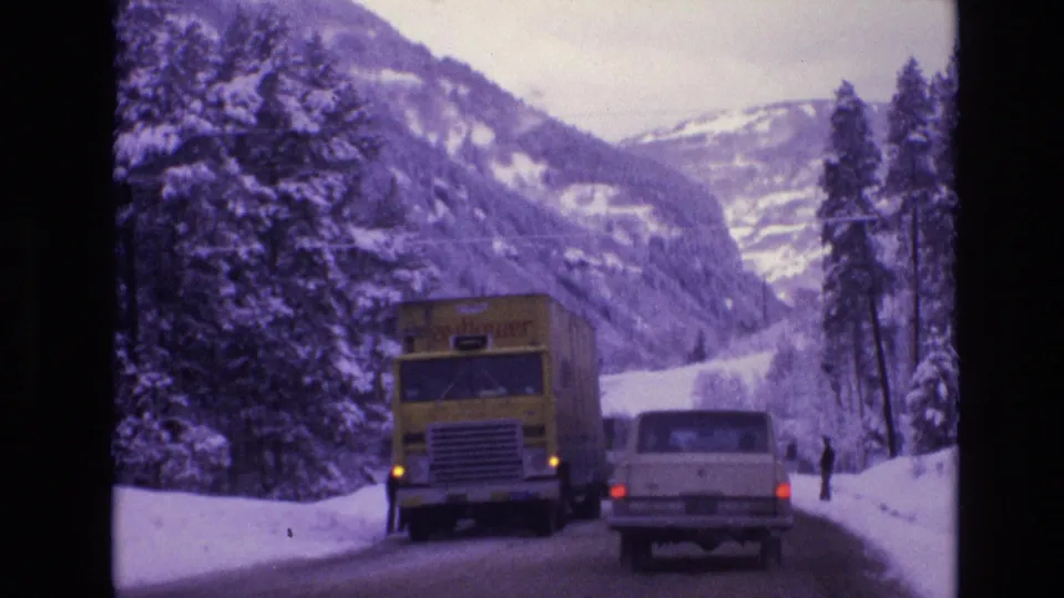 1974 Vail, Colorado frame showing a yellow truck, a white station wagon, a distant standing person, snowy evergreens, and mountain slopes along the road.