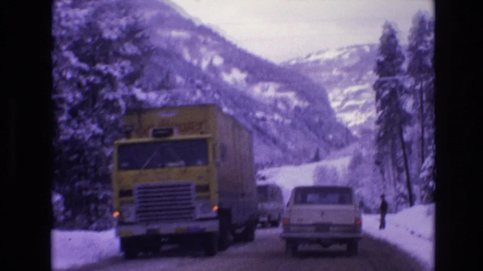 1974 Vail, Colorado home-movie frame with a yellow truck and white station wagon on a snow-lined road beneath forested mountain slopes.