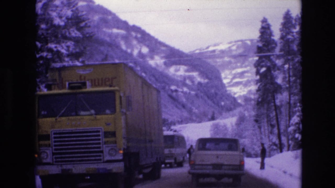 In supported 1974 Vail, Colorado footage, a yellow semi-truck with a trailer approaches on an icy road as cars follow between snowbanks and mountain slopes.