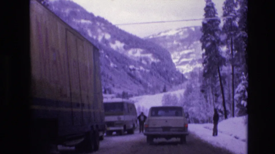The truck trailer fills the left side of the frame while a white van, a light-colored station wagon, and roadside pedestrians appear on a snow-lined Vail mountain road.