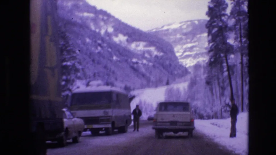 A partial yellow truck remains at the left edge as a white van, a light-colored station wagon, and two roadside figures occupy the snowy Vail road scene.