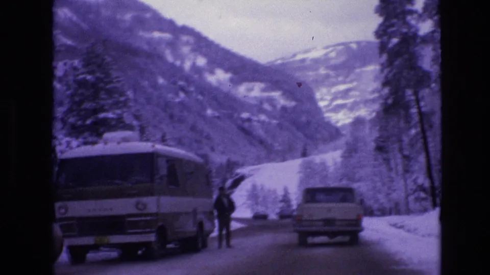 A white van or RV and a light-colored station wagon travel past a roadside person on a snowy Vail, Colorado mountain road with evergreen trees and winter slopes behind them.