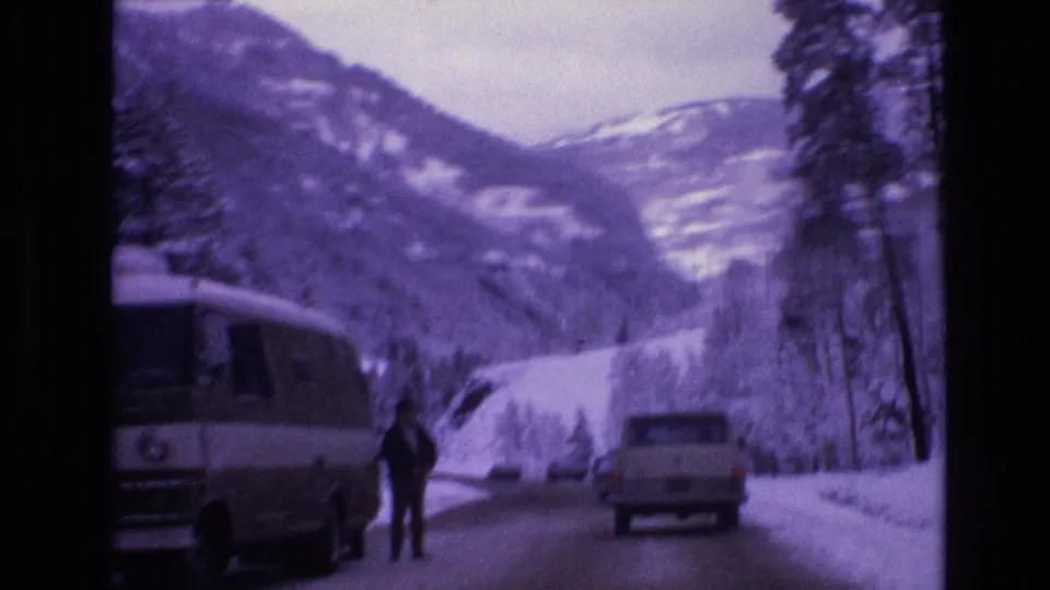 1974 Vail, Colorado 8mm frame showing a snowy mountain road with a camper van, a light pickup truck, a distant adult figure, pine trees, and snow-covered slopes.