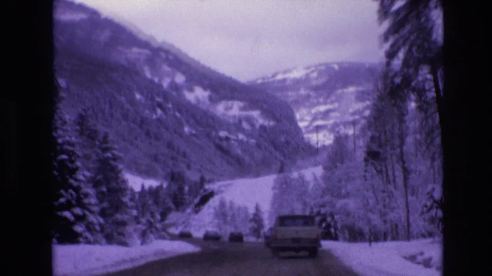 1974 Vail, Colorado 8mm frame showing a pickup truck on a snow-lined road beneath pine trees and layered mountain slopes.