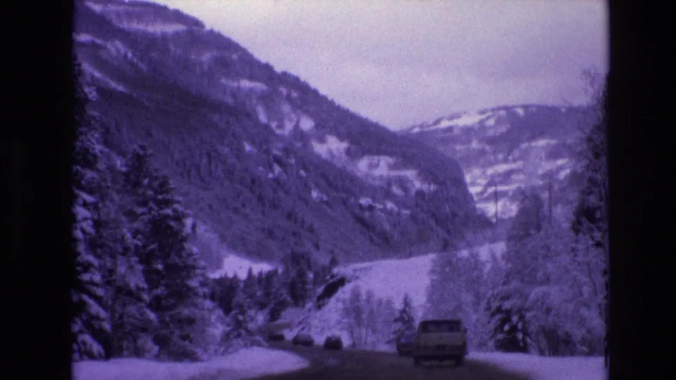 1974 Vail, Colorado 8mm frame showing a snow-lined mountain road with vehicles, evergreen trees, and steep winter slopes in the distance.