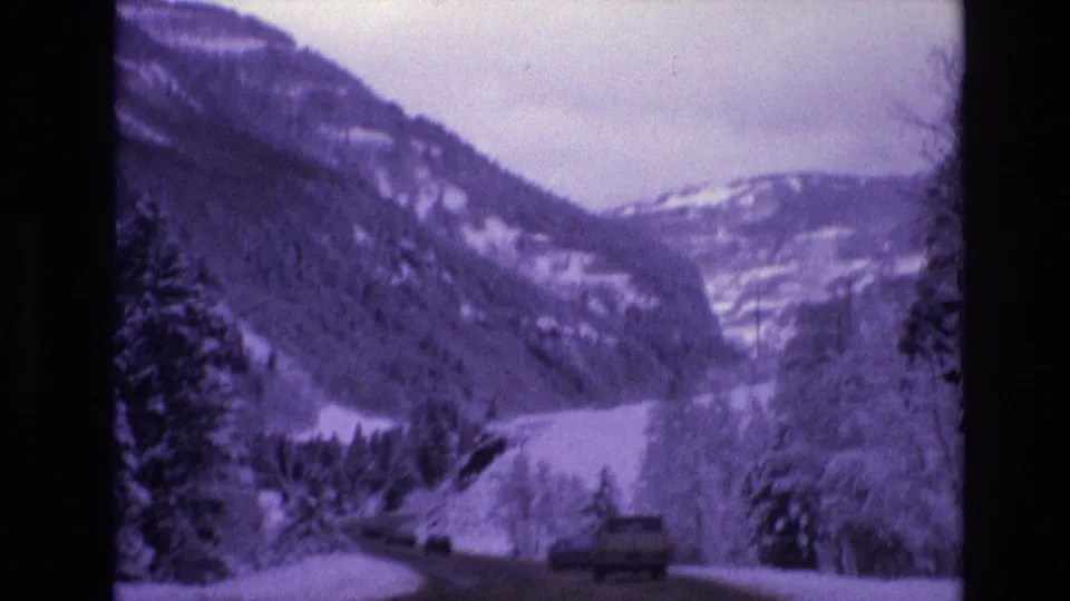 1974 Vail, Colorado frame of a vehicle on a winding winter road bordered by evergreens and snow-covered mountain terrain.