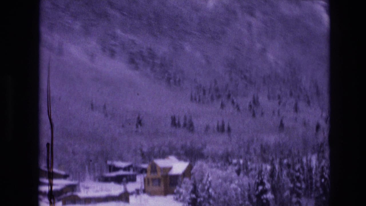 1974 Vail, Colorado home-movie frame showing a small snow-covered cabin near dark evergreens and a softly focused wooded mountainside.