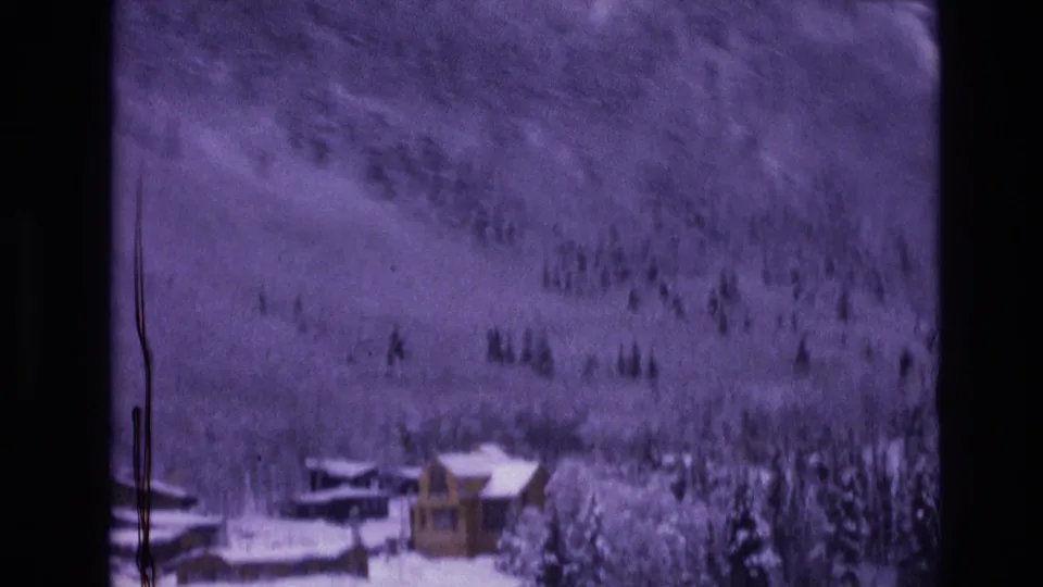 1974 Vail, Colorado home-movie frame showing a small snow-covered cabin near dark evergreens and a softly focused wooded mountainside.
