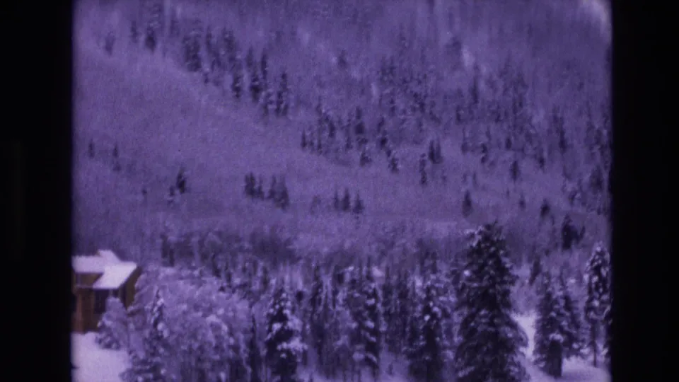 1974 Vail, Colorado home-movie still with a cabin partly cropped at the lower left and snow-laden evergreens rising toward a wooded slope.