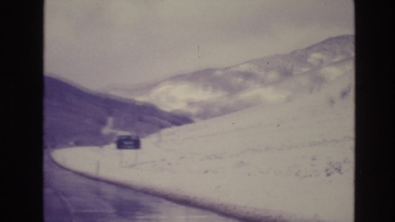 Snowy mountain road curving past a roadside sign and open slopes.
