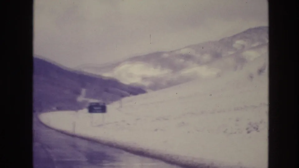 1974 Vail, Colorado road-travel frame showing a winding snow-lined mountain road, distant ridges, and a roadside sign viewed from a car.