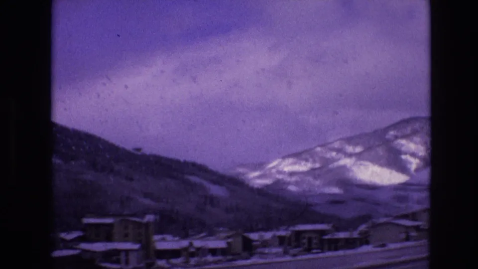 A moving-car view of low buildings and a road near snowy mountain slopes in Vail, Colorado in 1974 8mm footage.