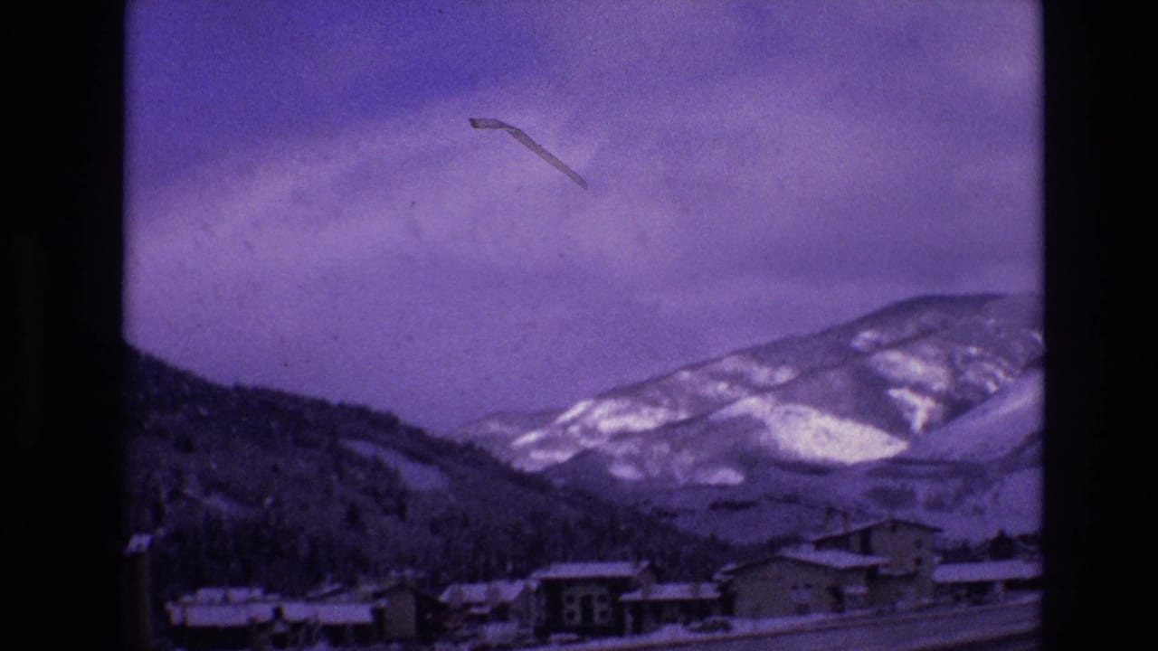 1974 Vail, Colorado home-movie frame showing snow-covered buildings in a valley beneath forested slopes and pale winter mountains under a purple-tinged sky.