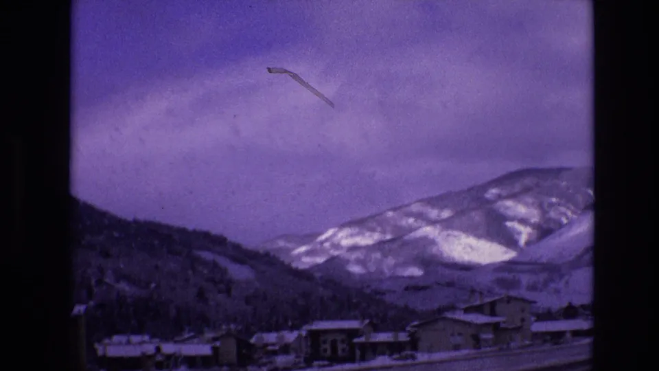 1974 Vail, Colorado home-movie frame showing snow-covered buildings in a valley beneath forested slopes and pale winter mountains under a purple-tinged sky.