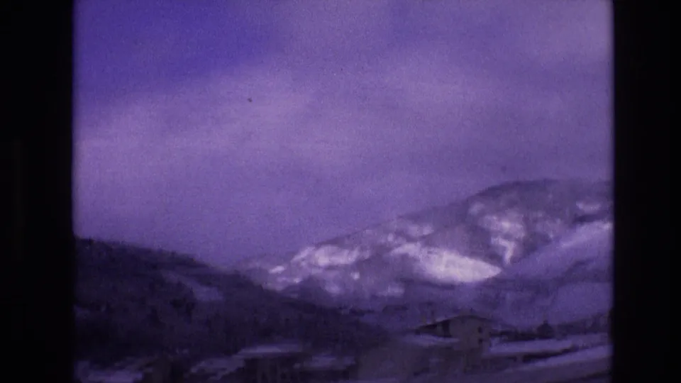 1974 Vail, Colorado frame from a moving home movie showing a snowy roadside, a lodge-like structure, wooded slopes, and distant snow-marked mountains.