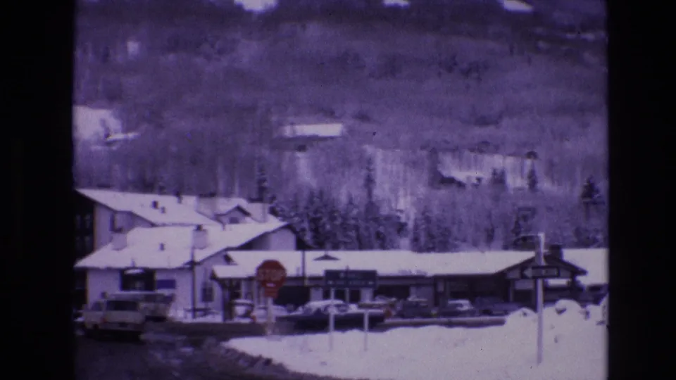 1974 Vail, Colorado home-movie frame showing roadside buildings, a stop sign, parked vehicles, deep snowbanks, leafless trees, and a wooded winter hillside.