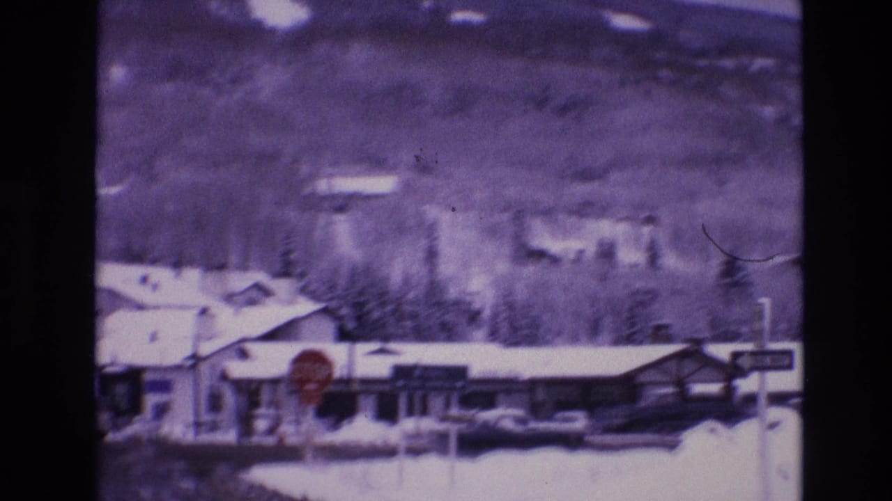 1974 Vail, Colorado 8mm frame showing a snowy roadside stop sign near low commercial buildings, passing cars, evergreen trees, and purple-toned mountains.