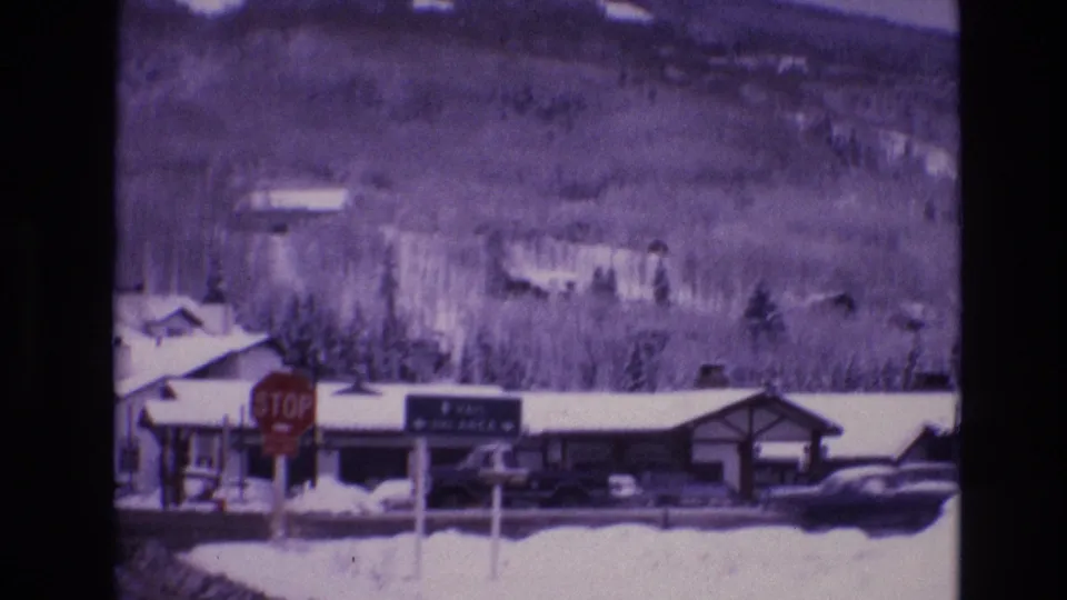 1974 Vail, Colorado frame with a red stop sign and green Vail Ski Area directional sign set against snow-covered buildings, cars, trees, and mountains.