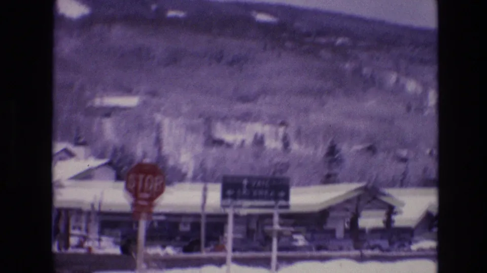 1974 Vail, Colorado 8mm frame showing a snow-lined street, red stop sign, Vail Ski Area wayfinding sign, lodge-like buildings, cars, and a wooded hillside.