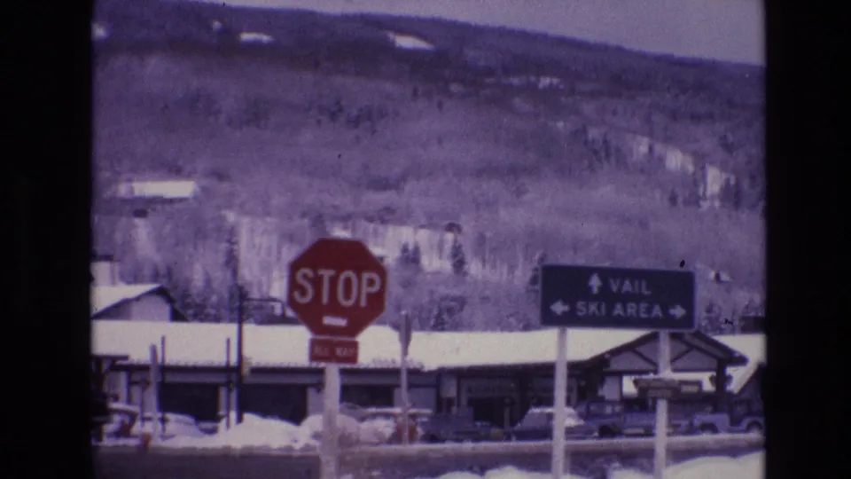 1974 Vail, Colorado frame with the stop sign and Vail Ski Area directional sign most prominent, backed by snowbanks, low buildings, cars, trees, and purple mountain slopes.