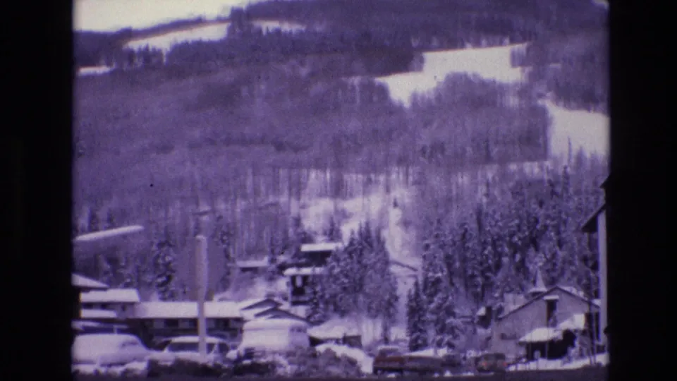 1974 Vail, Colorado roadside view with snowbanks, parked cars, small buildings, evergreen trees, and a forested mountain slope behind them.
