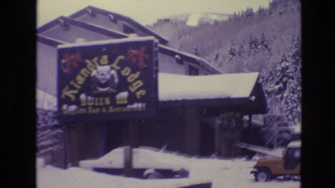 1974 Vail, Colorado frame with the Kiandra Lodge sign, snow-covered parked cars, lodge rooflines, evergreen trees, and a yellow Jeep-like vehicle at the edge of the lot.