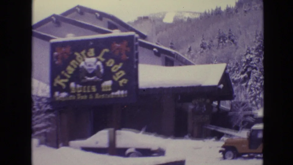 1974 Vail, Colorado frame with the Kiandra Lodge sign, snow-covered parked cars, lodge rooflines, evergreen trees, and a yellow Jeep-like vehicle at the edge of the lot.