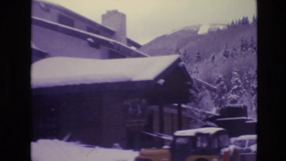 1974 Vail, Colorado home-movie frame showing a lodge roof heavy with snow, a chimney, parked vintage vehicles, trees, and the ski-slope hillside beyond.