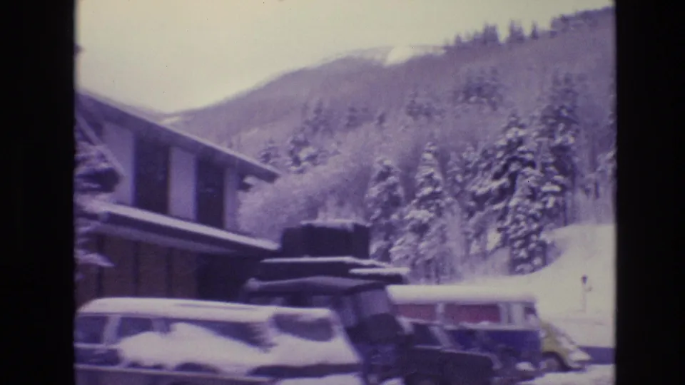 1974 Vail, Colorado frame showing snow-covered lodge buildings, several parked vintage cars, evergreen trees, and a winter mountain slope behind the resort area.